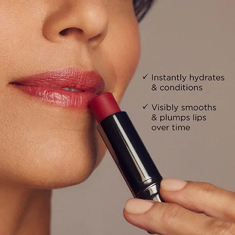 A person applies a reddish-pink lip balm to their lips, which are already hydrated, with text that reads: Instantly hydrates & conditions, Visibly smooths & plumps lips over time.
